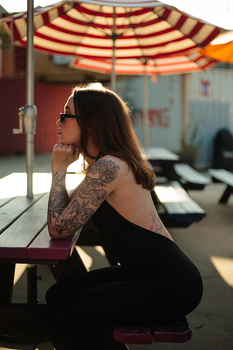 Woman with arm tattoos seated at an outdoor cafe under a striped umbrella.